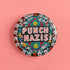 Round pinback metal button with an embroidered-style design on a black background. The center displays the phrase "PUNCH NAZIS" in bold, white stitched-textured lettering with red accents. Surrounding the text is an elaborate folk-inspired floral pattern, densely packed with embroidered-style flowers in shades of red, yellow, blue, pink, and white. The intricate design includes symmetrical lace-like stitching and decorative motifs, creating a rich, tapestry-like effect.